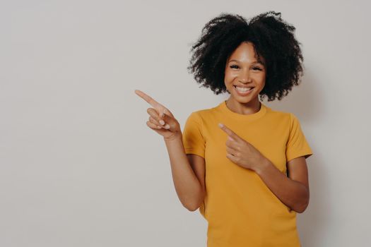 Look here. Studio portrait of beautiful cheerful dark skinned woman dressed in yellow tshirt pointing at left upper corner with forefingers, advertising item while standing against white background