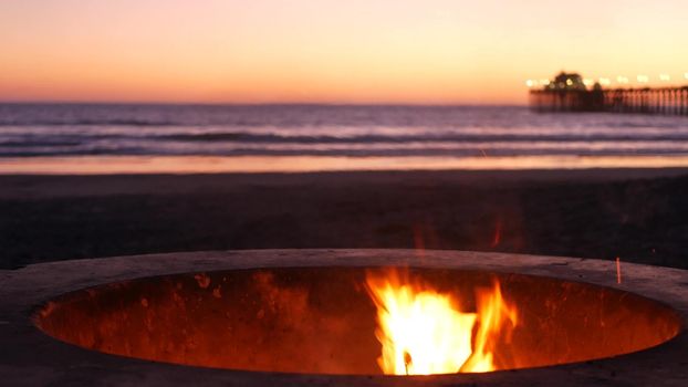 Campfire pit by Oceanside pier, California USA. Camp fire burning on ocean beach, bonfire flame in cement ring place for bbq, sea water waves. Romantic evening twilight sky, dusk after summer sunset.