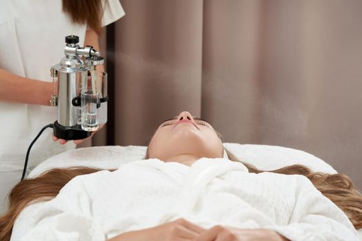 The procedure of steaming the skin of the face of a young woman before cleaning the skin in spa salon