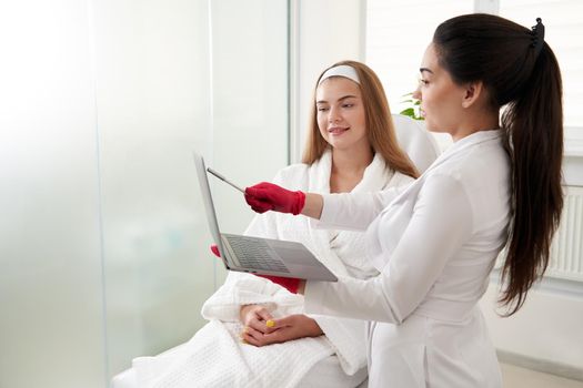 Female doctor working with patient while using laptop in clinic. Beauty doctor consults patient. Cosmetologist and beauty surgeon concept