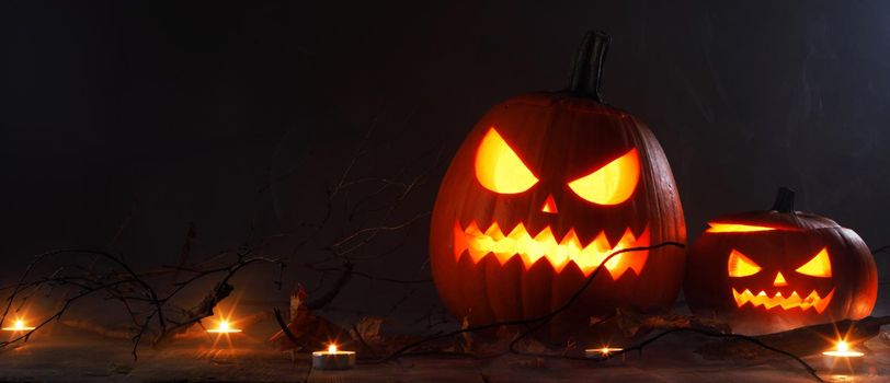 Halloween two luminous evil scary pumpkins, jack-lantern, with candles, leaves and eerie branches on wooden dark background