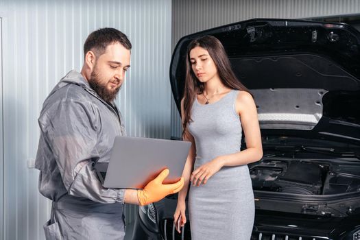 Car mechanic communicating with a customer while using laptop and examining vehicle breakdown at auto repair shop