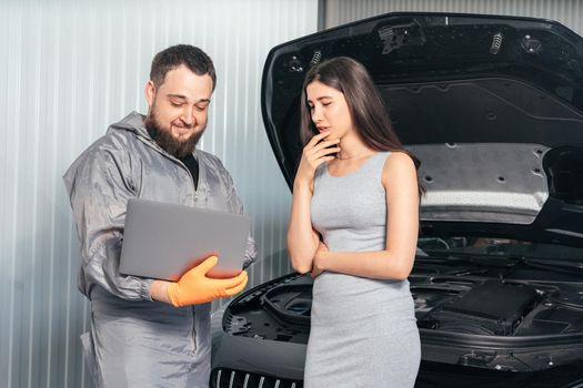 Car mechanic communicating with a customer while using laptop and examining vehicle breakdown at auto repair shop