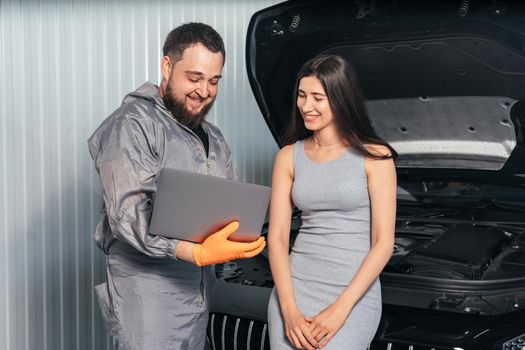 Car mechanic communicating with a customer while using laptop and examining vehicle breakdown at auto repair shop