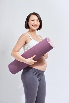 Joyful pregnant woman holding yoga mat smiling to camera posing over white background. Pregnant lifestyle and fitness.