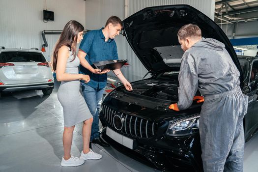 Car mechanic and customer stand next to the serviced car. The car had an annual checkup and stands in the garage with the hood open