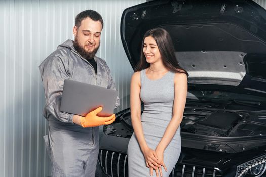 Car mechanic communicating with a customer while using laptop and examining vehicle breakdown at auto repair shop