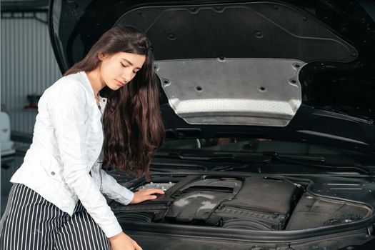 Young woman standing in front of the car with opened hood looking under a car hood
