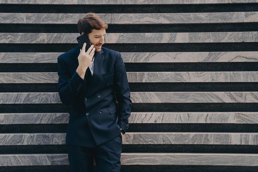 Businessman in elegant black clothes phones to partner focused down keeps smartphone near ear poses near marble wall copy space for your advertisement. People technology and bussines concept