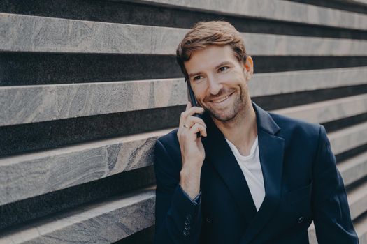 Handsome smiling man in stylish blazer and white t-shirt speaking on smartphone outside, smiling office worker having pleasant conversation on mobile phone with boss about last financial reports