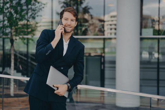 Confident young bearded businessman dressed in formal clothes making business call, standing outside of glass building with laptop in relaxed pose while talking on mobile phone with colleagues