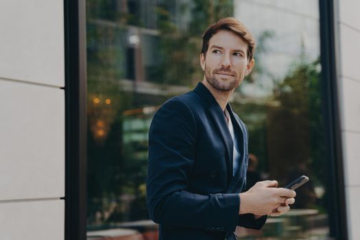 Successful handsome businessman with stubble messaging his employer about project details, holding smartphone in his hands while standing next to building with big window reflecting urban landscape