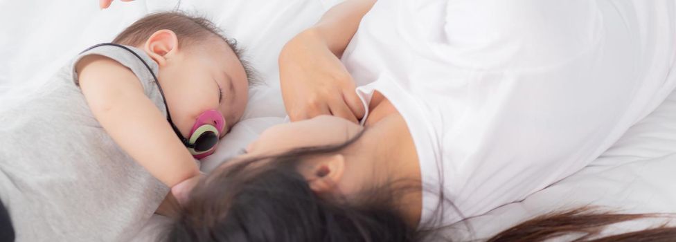 Young asian mother touch head of little baby girl with tender on bed in the bedroom, mom love newborn and care, woman with expression with child together, parent and daughter, family concept.