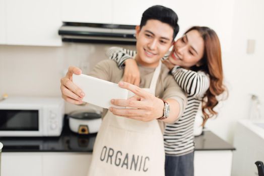 Happy asian couple smiling and cooking in the kitchen doing selfie using a smart phone