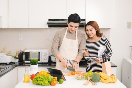 Young asian couple chopping vegetable and using digital tablet in the kitchen