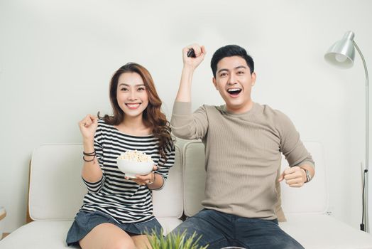 Excited young asian couple watching TV and eating popcorn