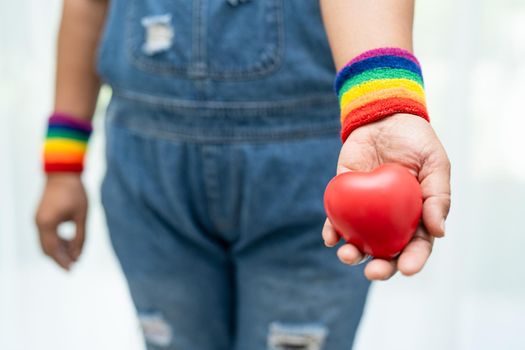 Asian lady wearing rainbow flag wristbands and hold red heart, symbol of LGBT pride month celebrate annual in June social of gay, lesbian, bisexual, transgender, human rights.