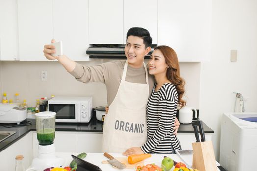 Happy asian couple smiling and cooking in the kitchen doing selfie using a smart phone