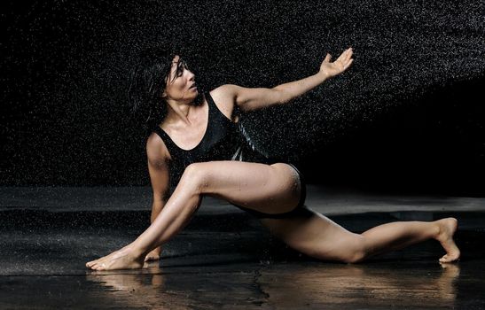 woman with black hair lies on the ground under raindrops on a black background. Woman dressed in black bodysuit