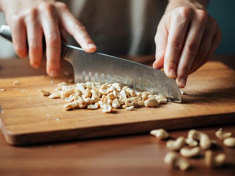 Female hands chopping cashew. Close up shot of process crushing raw unroasted cashew with large knife on wooden cutting board. Shallow DOF. Selective focus