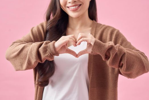 smiling young asian woman showing heart gesture with two hands and looking at camera isolated over pink background