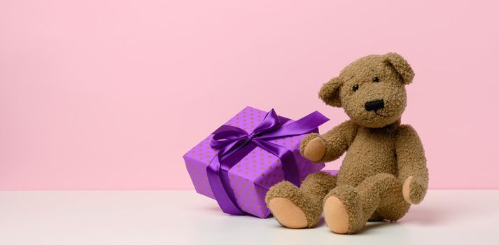 cute brown teddy bear holding a box wrapped in paper and red silk ribbon on white table. Prize and congratulations, pink background, copy space