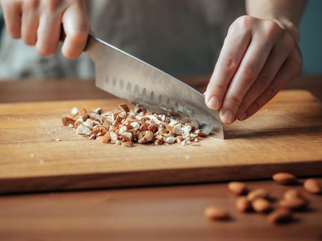 Female hands chopping almonds. Close up shot of process crushing raw unroasted almonds with large knife on wooden cutting board. Shallow DOF. Selective focus. Copy space
