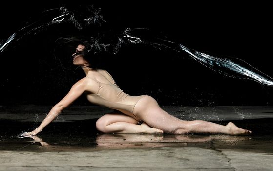 young woman with black long hair lies on the ground under raindrops on a black background. Woman dressed in beige bodysuit