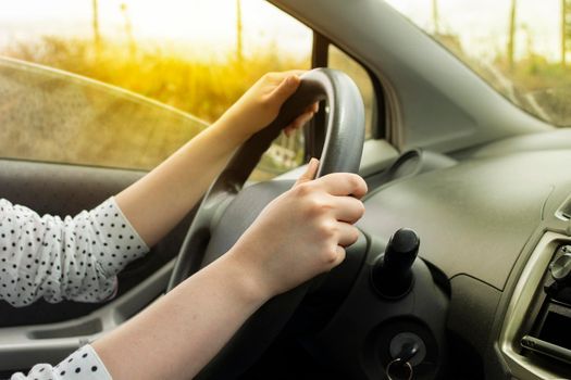 Close-up to the hands of a white woman on the steering wheel of a modern car with black upholstery in motion at sunset