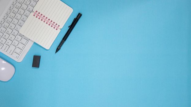 Office table desk with set of colorful supplies, blue blank note pad, cup, pen, pc, crumpled paper, flower on blue background. Top view and copy space for text