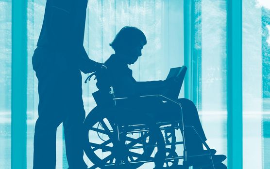 Elderly woman reading a book on wheelchair with her son take care.