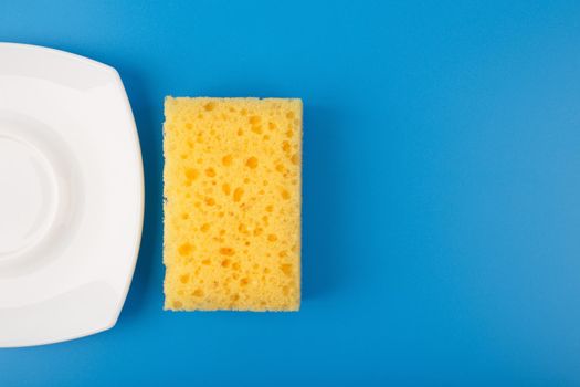 Dishwashing minimal creative concept. Close up of white shiny clean plate and yellow cleaning sponge against blue background with copy space. 