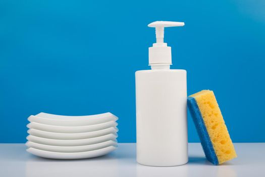 Colorful dishwashing concept, creative composition with liquid detergent in white plastic bottle with dispenser, yellow cleaning sponge and pile of clean plates against blue background