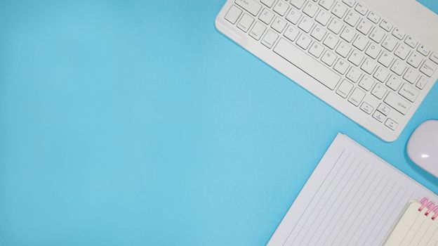 Office table desk with set of colorful supplies, blue blank note pad, cup, pen, pc, crumpled paper, flower on blue background. Top view and copy space for text