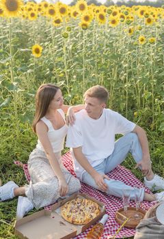 Leisure time. Young teenage couple picnic on sunflower field in sunset. Eating pizza and drinking champagne