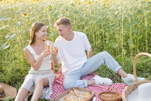 Young teenage couple picnic on sunflower field in sunset drinking champagne