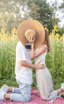 Romantic date. Fun and leisure. Young happy couple kissing on a picnic blanket, covering their faces with a summer hat