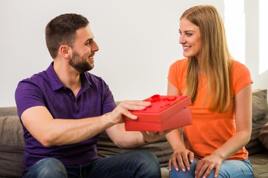 Husband giving present to his wife while they sitting on sofa at their home.