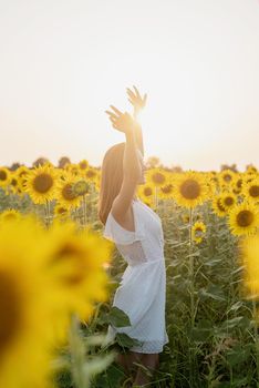 Freedom concept. Autumn nature. Young woman in white dress walking between sunflowers on a field in sunset raising her arms