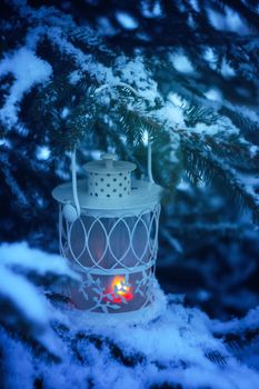 Decorative Christmas lantern with burning candle hanging on snow-covered fir- tree branch in a winter park. New year festive card, poster, postcard design.