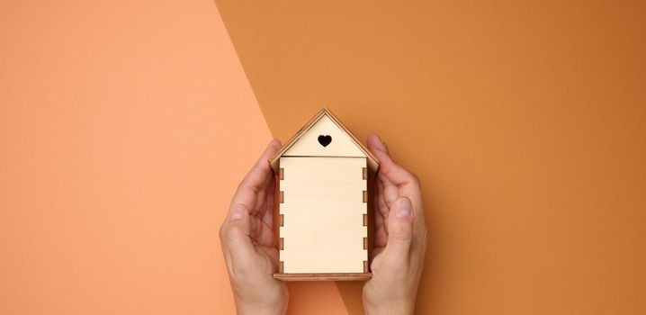 female hands folded to each other over a wooden miniature model house on a brown background. Real estate insurance concept, environmental protection, family happiness