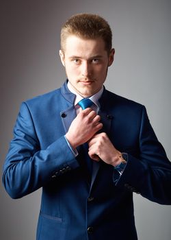 Handsome and self-confident. Handsome young man in formalwear adjusting his necktie and looking at camera while standing against grey background