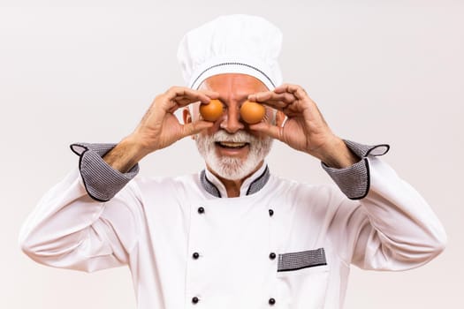 Image of happy senior chef covering eyes with eggs on gray background.