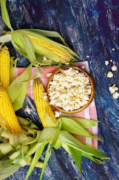Tasty popcorn in a bowl on the table. Ears of fresh corn on a dark background. Vertical photo