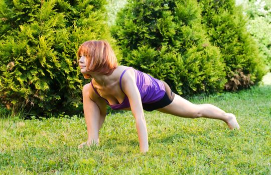 Beautiful red woman doing fitness or yoga exercises outdoors in a green park