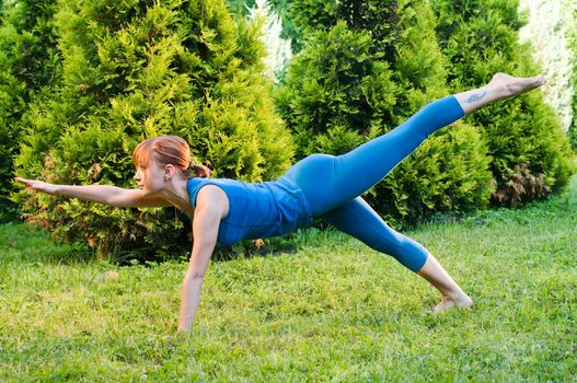 Beautiful red woman doing fitness or yoga exercises outdoors in a green park