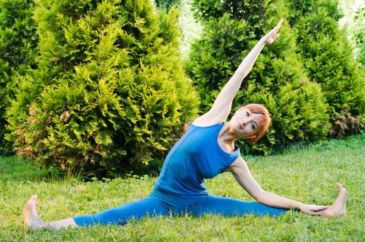 Beautiful red woman doing fitness or yoga exercises outdoors in a green park