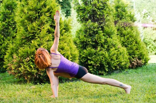 Beautiful red woman doing fitness or yoga exercises outdoors in a green park