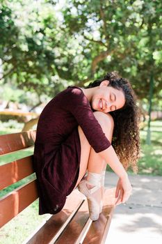 Young beautiful ballerina in black dress sitting on a bench in a park with her eyes closed and smiling