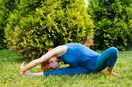 Beautiful red woman doing fitness or yoga exercises outdoors in a green park